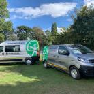 Two DEFRA silver electric vans parked on grass in a clearing within a wooded area.