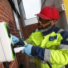 Man wearing a high visibility jacket and red hat insert a charging plug into a wall-mounted charging unit.