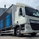 A white DAF semi truck with a blue-adorned curtain-side trailer parked beside industrial buildings under a clear sky.
