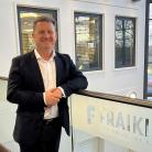  Generate by AI Assist  A person smiling in a business casual outfit, standing by a railing with a Fraikin sign, which reads "You Drive, We Care." The background shows a window overlooking a parking area.