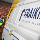 A person in safety gear is charging an electric vehicle beside a building. The van is white and yellow with the name "FRAIKIN" and the slogan "YOU DRIVE, WE CARE" displayed on its side.