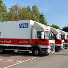 A row of NHS blood donation trucks parked in a lot. The trucks are white with a red section and a logo saying "Please give blood" alongside contact information. Trees and a clear sky are visible in the background.
