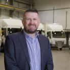 Man wearing a business suit stood in a warehouse in front of a line-up of various trucks