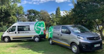 Two DEFRA silver electric vans parked on grass in a clearing within a wooded area.