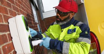 Man wearing a high visibility jacket and red hat insert a charging plug into a wall-mounted charging unit.