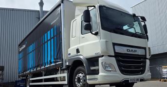 A white DAF semi truck with a blue-adorned curtain-side trailer parked beside industrial buildings under a clear sky.