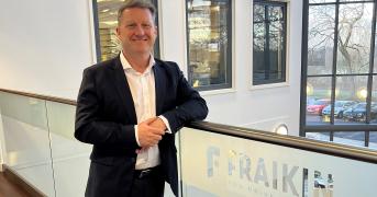  Generate by AI Assist  A person smiling in a business casual outfit, standing by a railing with a Fraikin sign, which reads "You Drive, We Care." The background shows a window overlooking a parking area.