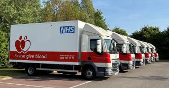 A row of NHS blood donation trucks parked in a lot. The trucks are white with a red section and a logo saying "Please give blood" alongside contact information. Trees and a clear sky are visible in the background.