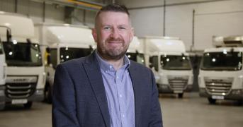 Man wearing a business suit stood in a warehouse in front of a line-up of various trucks