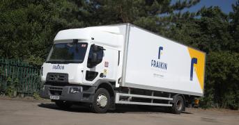 A Fraikin delivery truck is parked on a paved surface, surrounded by lush green trees and a blue sky. The truck bears the Fraikin logo and branding on its side.