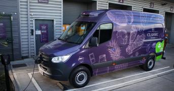 A purple delivery van is parked at an electric vehicle charging station outside a warehouse. The van features images of various breads and the word "Classic" on its side. The background shows a textured metal wall with numbered loading bays and a sign labeled "Driver's Entrance.