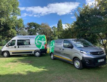 Two DEFRA silver electric vans parked on grass in a clearing within a wooded area.