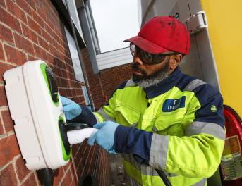 Man wearing a high visibility jacket and red hat insert a charging plug into a wall-mounted charging unit.