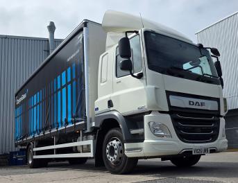 A white DAF semi truck with a blue-adorned curtain-side trailer parked beside industrial buildings under a clear sky.