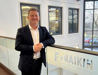  Generate by AI Assist  A person smiling in a business casual outfit, standing by a railing with a Fraikin sign, which reads "You Drive, We Care." The background shows a window overlooking a parking area.