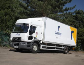 A Fraikin delivery truck is parked on a paved surface, surrounded by lush green trees and a blue sky. The truck bears the Fraikin logo and branding on its side.