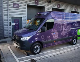 A purple delivery van is parked at an electric vehicle charging station outside a warehouse. The van features images of various breads and the word "Classic" on its side. The background shows a textured metal wall with numbered loading bays and a sign labeled "Driver's Entrance.