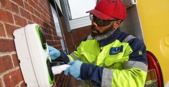 Man wearing a high visibility jacket and red hat insert a charging plug into a wall-mounted charging unit.