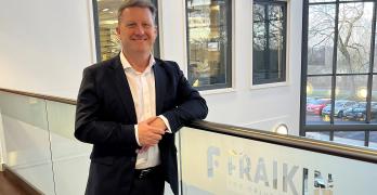  Generate by AI Assist  A person smiling in a business casual outfit, standing by a railing with a Fraikin sign, which reads "You Drive, We Care." The background shows a window overlooking a parking area.