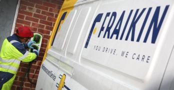 A person in safety gear is charging an electric vehicle beside a building. The van is white and yellow with the name "FRAIKIN" and the slogan "YOU DRIVE, WE CARE" displayed on its side.