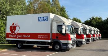 A row of NHS blood donation trucks parked in a lot. The trucks are white with a red section and a logo saying "Please give blood" alongside contact information. Trees and a clear sky are visible in the background.