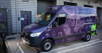 A purple delivery van is parked at an electric vehicle charging station outside a warehouse. The van features images of various breads and the word "Classic" on its side. The background shows a textured metal wall with numbered loading bays and a sign labeled "Driver's Entrance.